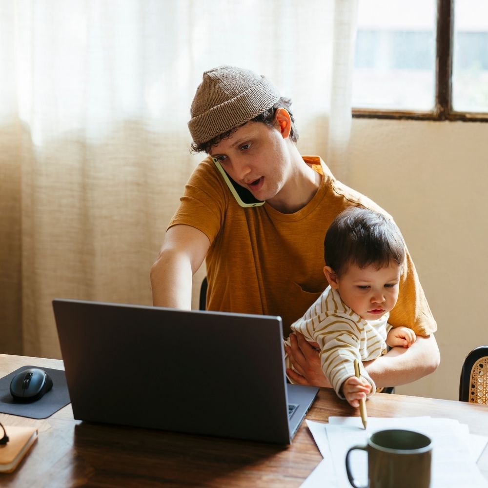 Caregiver on the phone holding a baby and using a computer, pursuing available resources